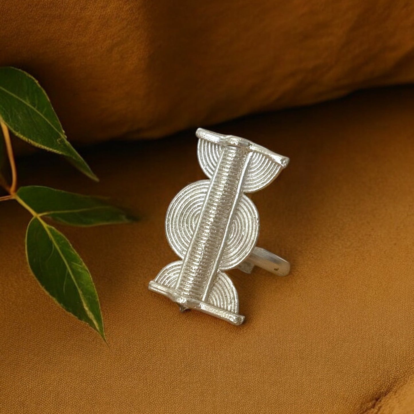 A silver-toned brass ring with geometric design, displayed on a brown textured surface with a green leaf in the background.